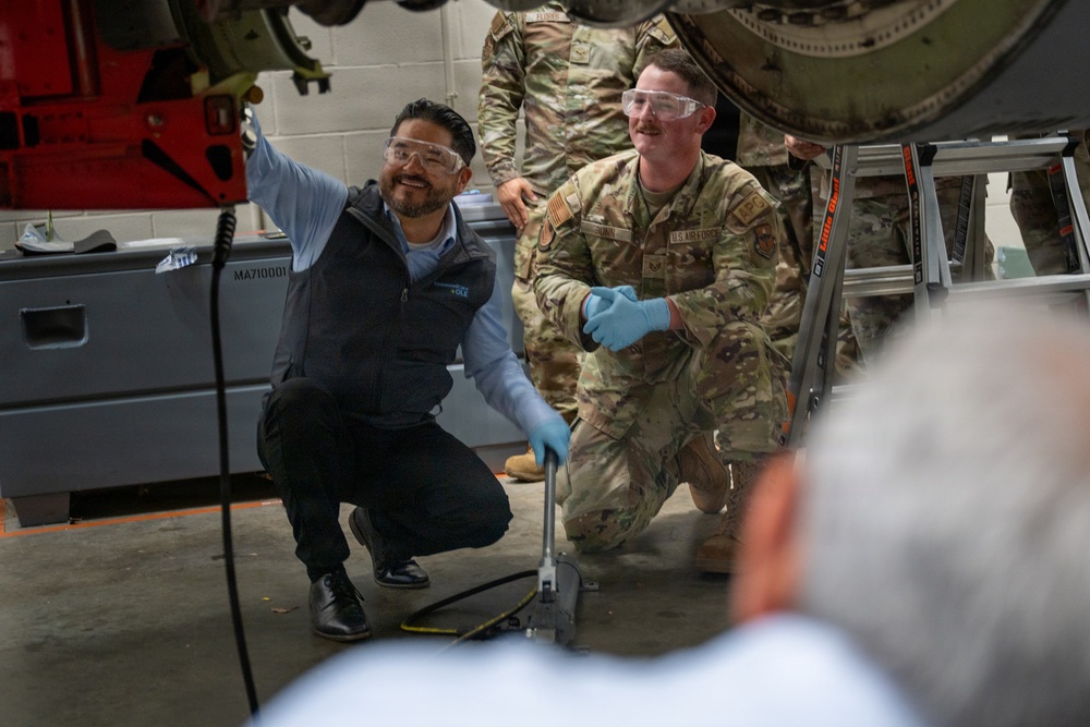 A civilian contractor and a U.S. Air Force service member are crouched together in a maintenance hangar, working on what appears to be aircraft or military vehicle equipment. Both individuals are wearing safety goggles and blue nitrile gloves, suggesting a hands-on technical maintenance or inspection task. Additional uniformed personnel are visible in the background.