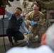A civilian contractor and a U.S. Air Force service member are crouched together in a maintenance hangar, working on what appears to be aircraft or military vehicle equipment. Both individuals are wearing safety goggles and blue nitrile gloves, suggesting a hands-on technical maintenance or inspection task. Additional uniformed personnel are visible in the background.