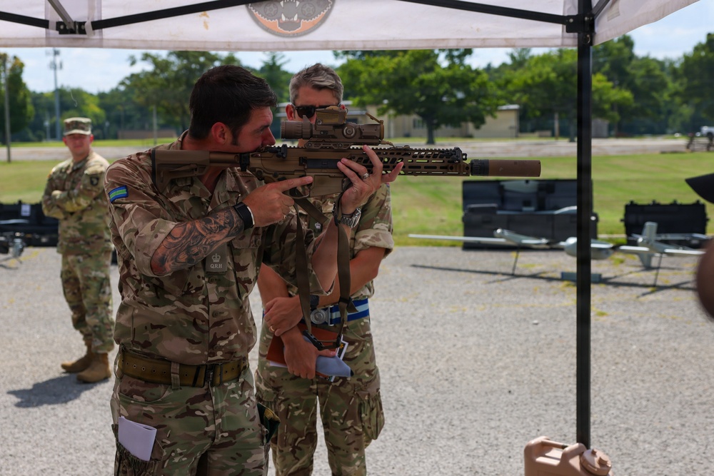 Military personnel are gathered under a canopy tent at what appears to be a weapons demonstration or evaluation event. A soldier wearing British Army MultiCam uniform with 'QRH' (Queen's Royal Hussars) insignia is aiming a modern rifle with optics and suppressor, while a colleague observes closely. A US Army soldier in OCP uniform is visible in the background.