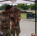 Military personnel are gathered under a canopy tent at what appears to be a weapons demonstration or evaluation event. A soldier wearing British Army MultiCam uniform with 'QRH' (Queen's Royal Hussars) insignia is aiming a modern rifle with optics and suppressor, while a colleague observes closely. A US Army soldier in OCP uniform is visible in the background.