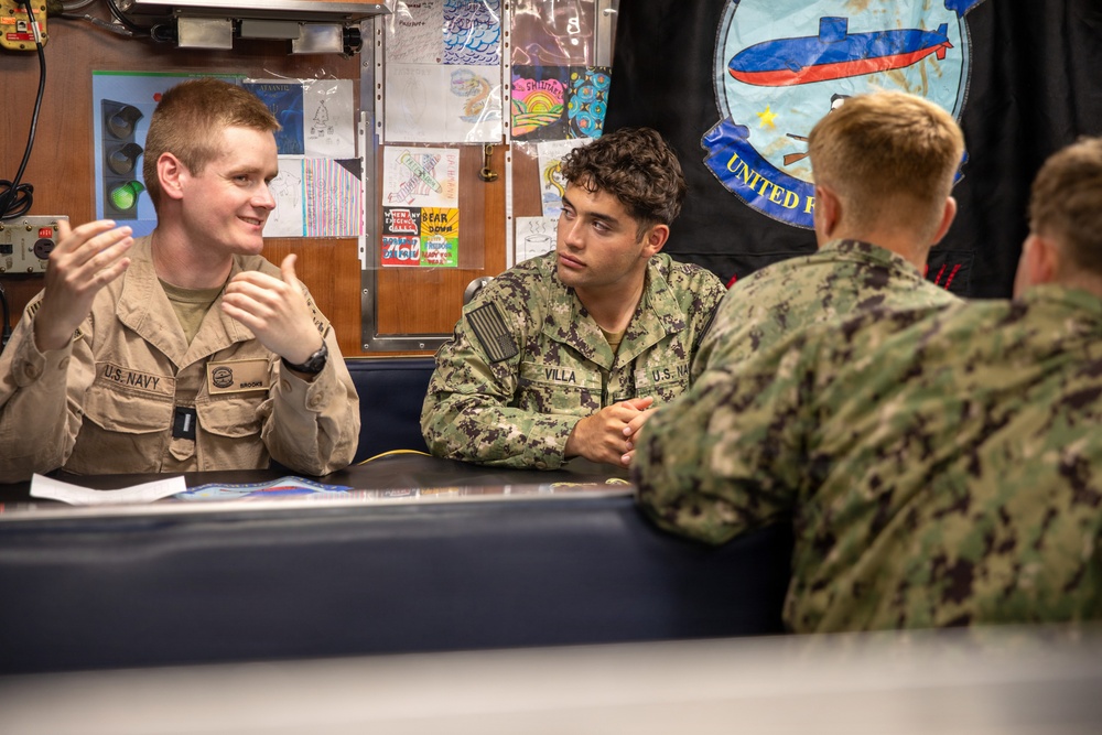 Three U.S. Navy personnel are seated at a table in what appears to be an onboard compartment or classroom, engaged in conversation. Two sailors face the camera while a third has their back to the viewer. Children's artwork and a submarine-themed banner are visible in the background.