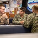 Three U.S. Navy personnel are seated at a table in what appears to be an onboard compartment or classroom, engaged in conversation. Two sailors face the camera while a third has their back to the viewer. Children's artwork and a submarine-themed banner are visible in the background.