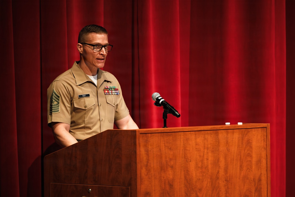 A military service member in tan/khaki uniform is speaking at a wooden podium on a stage with a red curtain backdrop. The individual wears multiple service ribbons, rank insignia, and a name tag reading 'RAY'. The setting appears to be a formal military ceremony or briefing.