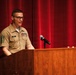 A military service member in tan/khaki uniform is speaking at a wooden podium on a stage with a red curtain backdrop. The individual wears multiple service ribbons, rank insignia, and a name tag reading 'RAY'. The setting appears to be a formal military ceremony or briefing.