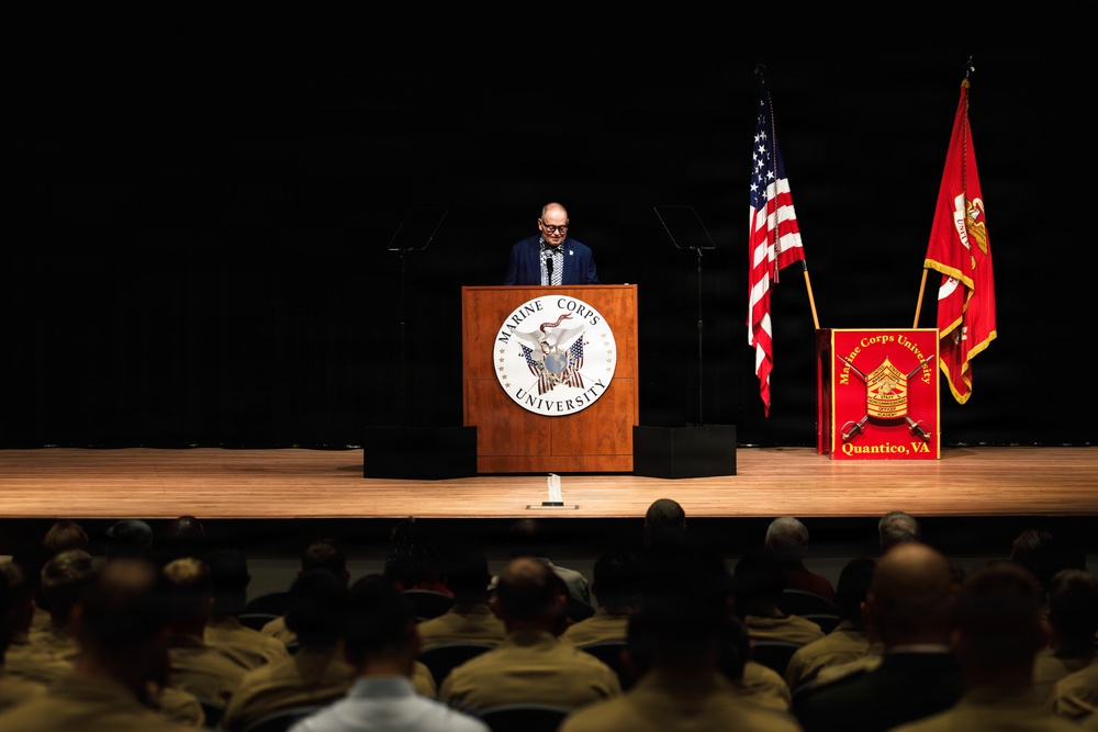 A civilian speaker addresses an audience of uniformed military personnel at Marine Corps University in Quantico, Virginia. The speaker stands at a wooden podium bearing the Marine Corps University seal, flanked by the United States flag and the Marine Corps University flag. The auditorium is filled with what appear to be Marines in tan/khaki uniforms seated in rows.