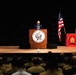 A civilian speaker addresses an audience of uniformed military personnel at Marine Corps University in Quantico, Virginia. The speaker stands at a wooden podium bearing the Marine Corps University seal, flanked by the United States flag and the Marine Corps University flag. The auditorium is filled with what appear to be Marines in tan/khaki uniforms seated in rows.