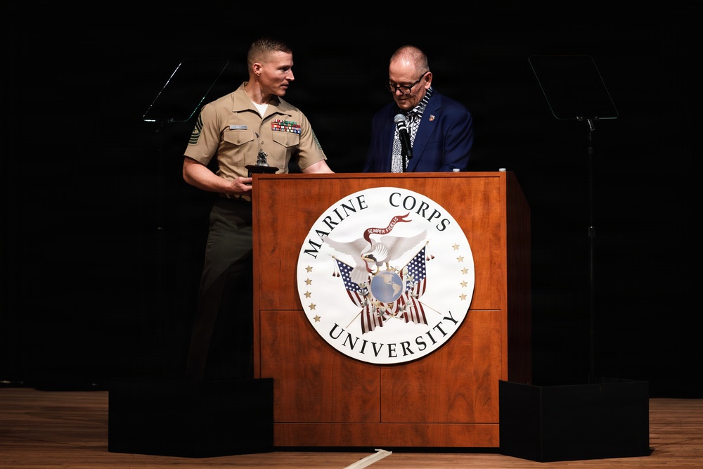 Two individuals stand at a Marine Corps University podium during what appears to be a formal ceremony or presentation. A uniformed Marine in service uniform with visible rank insignia and service ribbons stands alongside a civilian man in a blue blazer and patterned tie. The Marine appears to be holding an award or plaque.