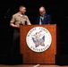 Two individuals stand at a Marine Corps University podium during what appears to be a formal ceremony or presentation. A uniformed Marine in service uniform with visible rank insignia and service ribbons stands alongside a civilian man in a blue blazer and patterned tie. The Marine appears to be holding an award or plaque.
