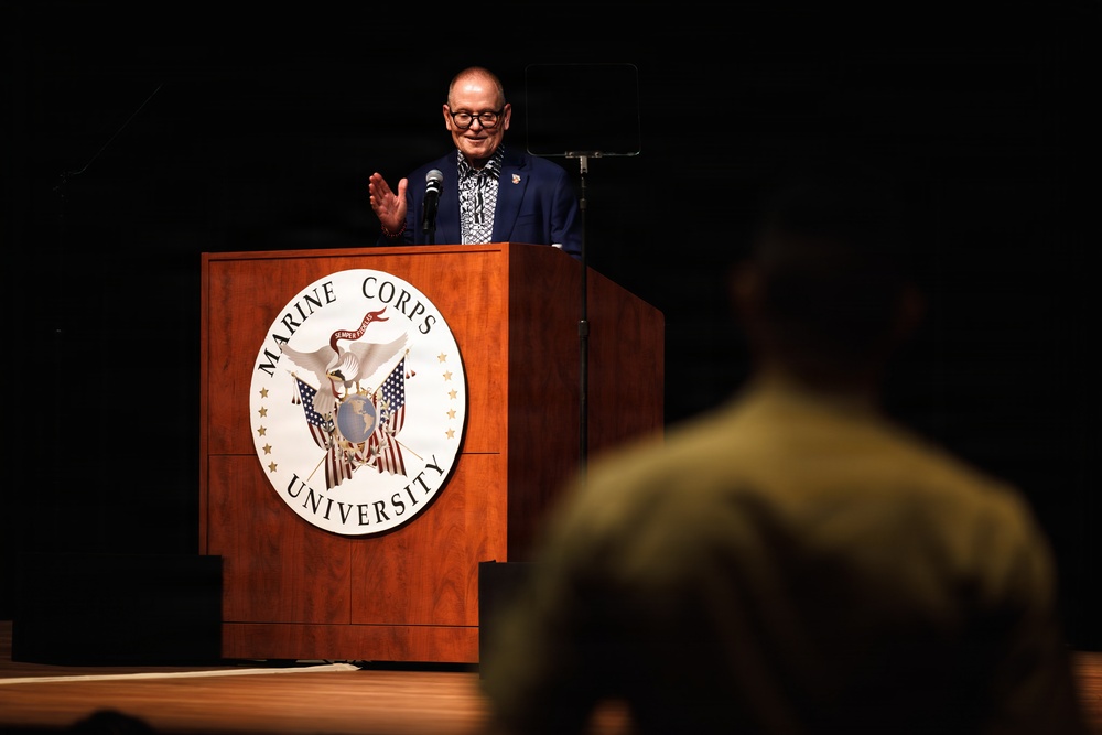A civilian speaker addresses an audience from a wooden podium bearing the Marine Corps University seal. The speaker, wearing a blue blazer and patterned shirt, appears to be mid-speech with one hand raised in a gesturing motion. A partially visible audience member in what appears to be military attire is visible in the foreground.