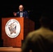 A civilian speaker addresses an audience from a wooden podium bearing the Marine Corps University seal. The speaker, wearing a blue blazer and patterned shirt, appears to be mid-speech with one hand raised in a gesturing motion. A partially visible audience member in what appears to be military attire is visible in the foreground.