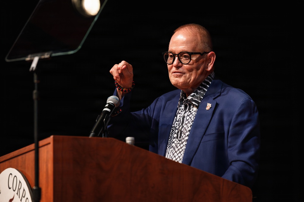 An older man with a shaved head and thick black-rimmed glasses is speaking at a wooden podium equipped with a microphone. He is wearing a blue blazer over a patterned black-and-white shirt and has a red beaded bracelet on his raised wrist. The podium bears partial text reading 'CORP...' suggesting an organizational or institutional event.