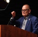 An older man with a shaved head and thick black-rimmed glasses is speaking at a wooden podium equipped with a microphone. He is wearing a blue blazer over a patterned black-and-white shirt and has a red beaded bracelet on his raised wrist. The podium bears partial text reading 'CORP...' suggesting an organizational or institutional event.