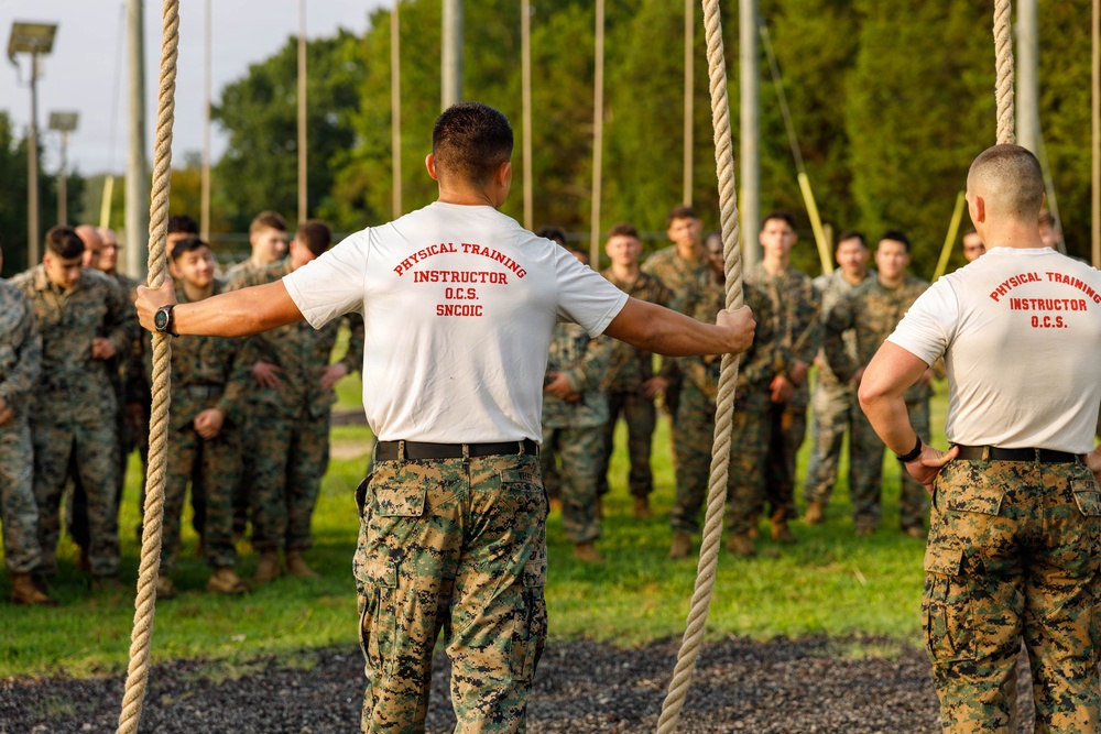 Two Physical Training Instructors from O.C.S. (Officer Candidate School) stand facing a large group of candidates at what appears to be a rope climb obstacle course. The instructors, wearing white PT shirts with 'Physical Training Instructor O.C.S. SNCOIC' printed in red, are positioned between hanging ropes while addressing the assembled candidates. The candidates are dressed in MARPAT (Marine pattern) camouflage uniforms and are standing in formation.