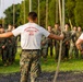 Two Physical Training Instructors from O.C.S. (Officer Candidate School) stand facing a large group of candidates at what appears to be a rope climb obstacle course. The instructors, wearing white PT shirts with 'Physical Training Instructor O.C.S. SNCOIC' printed in red, are positioned between hanging ropes while addressing the assembled candidates. The candidates are dressed in MARPAT (Marine pattern) camouflage uniforms and are standing in formation.