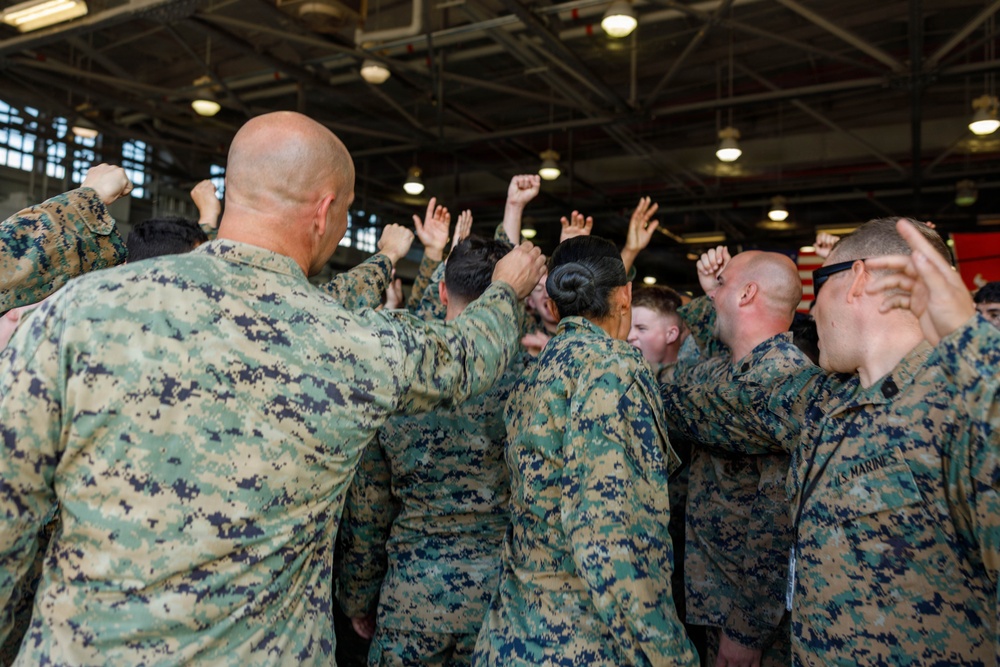 A group of U.S. Marines in MARPAT digital camouflage uniforms are gathered in what appears to be a hangar bay, raising their fists in a group motivational huddle. The scene suggests a unit rally, morale event, or pre-deployment ceremony. An American flag is partially visible in the background.
