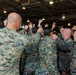 A group of U.S. Marines in MARPAT digital camouflage uniforms are gathered in what appears to be a hangar bay, raising their fists in a group motivational huddle. The scene suggests a unit rally, morale event, or pre-deployment ceremony. An American flag is partially visible in the background.