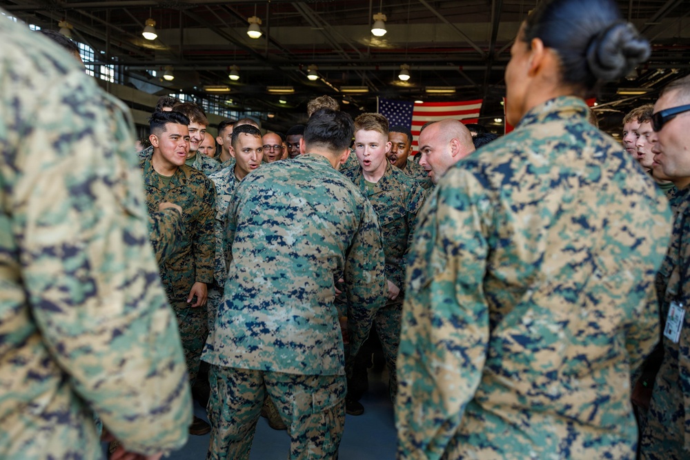 A group of U.S. Marines in MARPAT (Marine Pattern) digital camouflage uniforms are gathered in what appears to be a large indoor facility, possibly a hangar or gymnasium aboard a naval vessel or military installation. The personnel appear to be engaged in a team huddle or motivational gathering, with several individuals visibly shouting or chanting. An American flag is visible in the background.