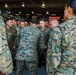 A group of U.S. Marines in MARPAT (Marine Pattern) digital camouflage uniforms are gathered in what appears to be a large indoor facility, possibly a hangar or gymnasium aboard a naval vessel or military installation. The personnel appear to be engaged in a team huddle or motivational gathering, with several individuals visibly shouting or chanting. An American flag is visible in the background.