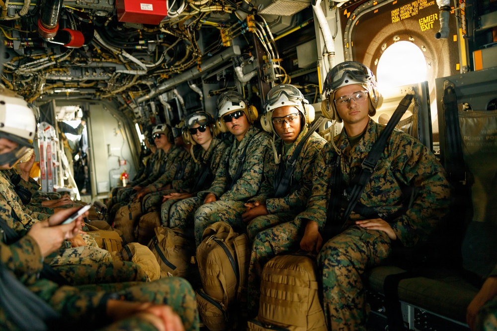 A group of military personnel in camouflage uniforms are seated inside what appears to be a military transport helicopter or aircraft. They are wearing flight helmets with ear protection and protective eyewear, with tactical gear and duffel bags at their feet. An emergency exit sign is visible on the right side of the aircraft interior.