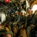 A group of military personnel in camouflage uniforms are seated inside what appears to be a military transport helicopter or aircraft. They are wearing flight helmets with ear protection and protective eyewear, with tactical gear and duffel bags at their feet. An emergency exit sign is visible on the right side of the aircraft interior.
