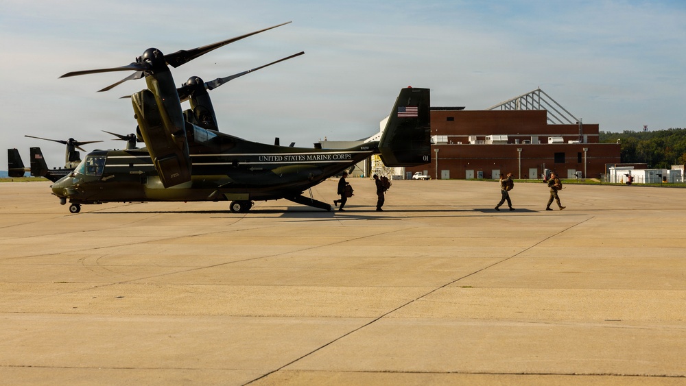 A United States Marine Corps MV-22 Osprey tiltrotor aircraft is parked on a tarmac, with personnel boarding or disembarking. The aircraft displays 'UNITED STATES MARINE CORPS' lettering and an American flag on its tail. A second helicopter is visible in the background to the left, and a large brick hangar building is visible on the right.