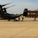 A United States Marine Corps MV-22 Osprey tiltrotor aircraft is parked on a tarmac, with personnel boarding or disembarking. The aircraft displays 'UNITED STATES MARINE CORPS' lettering and an American flag on its tail. A second helicopter is visible in the background to the left, and a large brick hangar building is visible on the right.