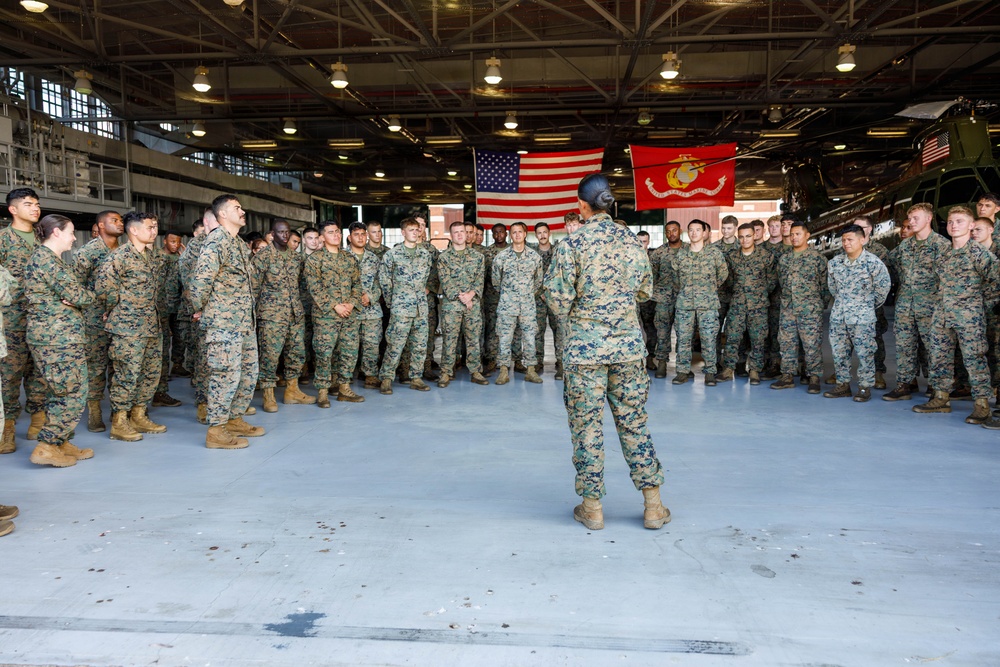 A military formation is assembled inside a large hangar facility, with one service member standing with their back to the camera addressing the assembled group. Two flags are prominently displayed in the background: the United States flag and the United States Marine Corps flag. The setting and uniforms indicate this is a U.S. Marine Corps unit briefing or formation.
