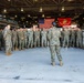 A military formation is assembled inside a large hangar facility, with one service member standing with their back to the camera addressing the assembled group. Two flags are prominently displayed in the background: the United States flag and the United States Marine Corps flag. The setting and uniforms indicate this is a U.S. Marine Corps unit briefing or formation.