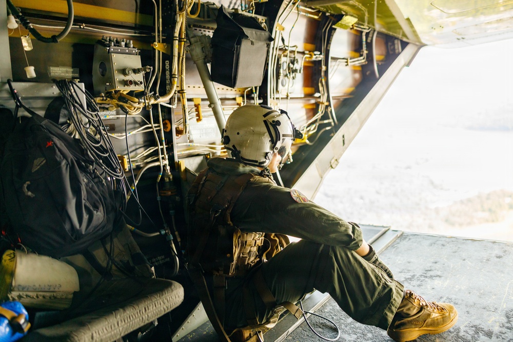 A military aircrew member wearing a flight helmet and olive drab flight suit is seated at the open rear ramp of a military transport aircraft, looking out over terrain below. The interior of the aircraft shows extensive wiring, control panels, and yellow-painted structural components typical of military cargo aircraft. The person appears to be performing observation or door guard duties during flight.