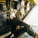 A military aircrew member wearing a flight helmet and olive drab flight suit is seated at the open rear ramp of a military transport aircraft, looking out over terrain below. The interior of the aircraft shows extensive wiring, control panels, and yellow-painted structural components typical of military cargo aircraft. The person appears to be performing observation or door guard duties during flight.