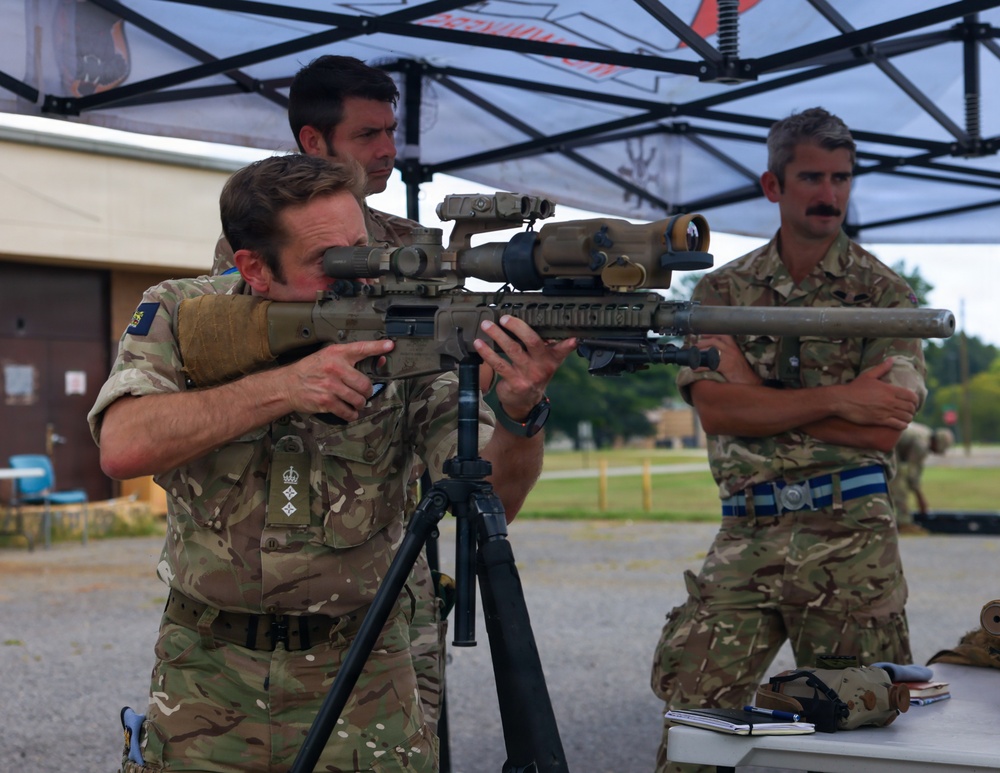 Three military personnel in MultiCam/MTP camouflage uniforms are gathered under a canopy tent at what appears to be a shooting range or military training event. One soldier is aiming a precision rifle mounted on a tripod, equipped with multiple optics including a large scope and red dot sight. The others observe, with one taking notes at a table.