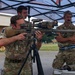 Three military personnel in MultiCam/MTP camouflage uniforms are gathered under a canopy tent at what appears to be a shooting range or military training event. One soldier is aiming a precision rifle mounted on a tripod, equipped with multiple optics including a large scope and red dot sight. The others observe, with one taking notes at a table.