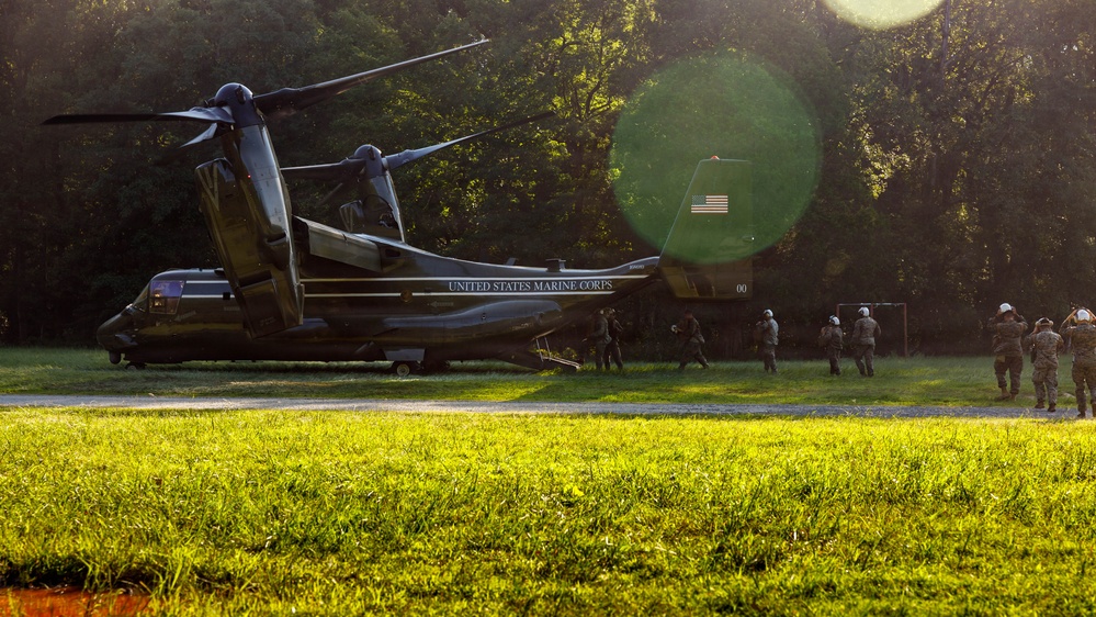A United States Marine Corps V-22 Osprey tiltrotor aircraft is landed on a grassy field surrounded by trees, with personnel boarding or disembarking via the rear ramp. The image is taken during golden hour lighting with visible lens flare, giving it a dramatic quality. An American flag is visible on the tail section of the aircraft.