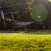 A United States Marine Corps V-22 Osprey tiltrotor aircraft is landed on a grassy field surrounded by trees, with personnel boarding or disembarking via the rear ramp. The image is taken during golden hour lighting with visible lens flare, giving it a dramatic quality. An American flag is visible on the tail section of the aircraft.