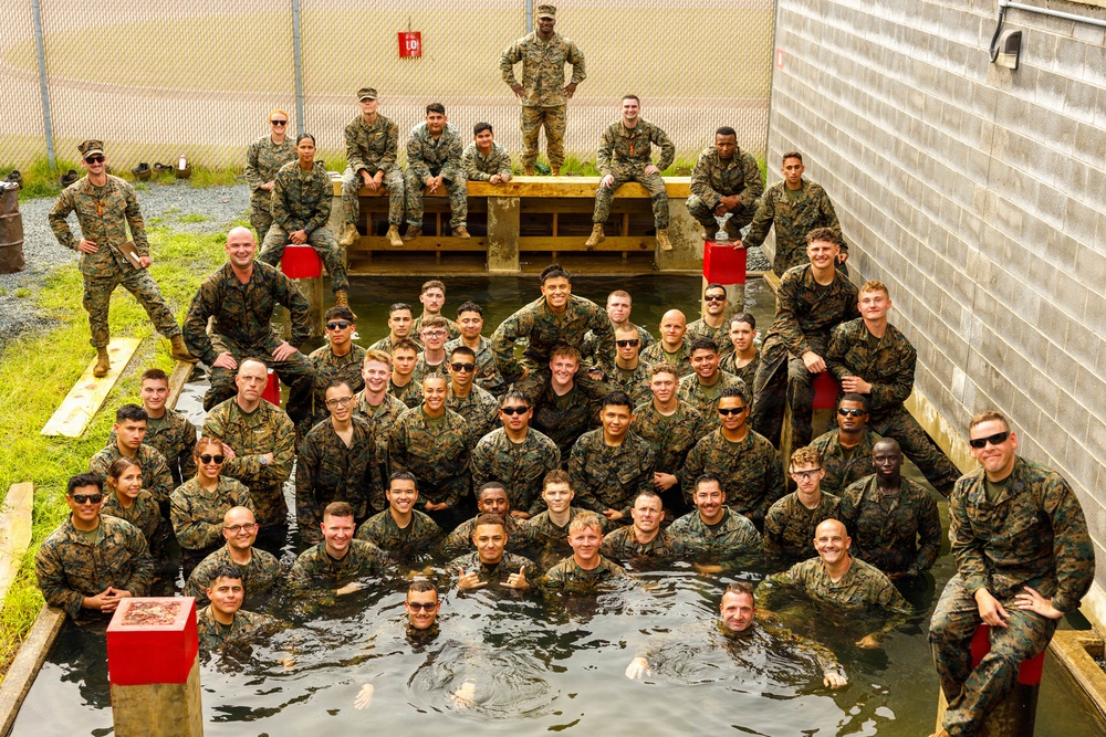 A large group of military personnel in camouflage uniforms pose for a group photo around and inside a water obstacle or training pool. The group appears to be celebrating or commemorating the completion of a water-based military training exercise. Personnel are arranged in tiered rows, with some standing on dry ground, some sitting on wooden structures, and others partially or fully submerged in the water.