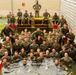 A large group of military personnel in camouflage uniforms pose for a group photo around and inside a water obstacle or training pool. The group appears to be celebrating or commemorating the completion of a water-based military training exercise. Personnel are arranged in tiered rows, with some standing on dry ground, some sitting on wooden structures, and others partially or fully submerged in the water.