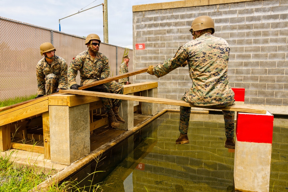 A group of military personnel in camouflage uniforms and helmets are engaged in what appears to be a team-building or obstacle course exercise involving a water obstacle. They are working with wooden planks to bridge or navigate across a water-filled channel. The setting appears to be a military training facility with concrete block walls and chain-link fencing.