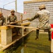 A group of military personnel in camouflage uniforms and helmets are engaged in what appears to be a team-building or obstacle course exercise involving a water obstacle. They are working with wooden planks to bridge or navigate across a water-filled channel. The setting appears to be a military training facility with concrete block walls and chain-link fencing.