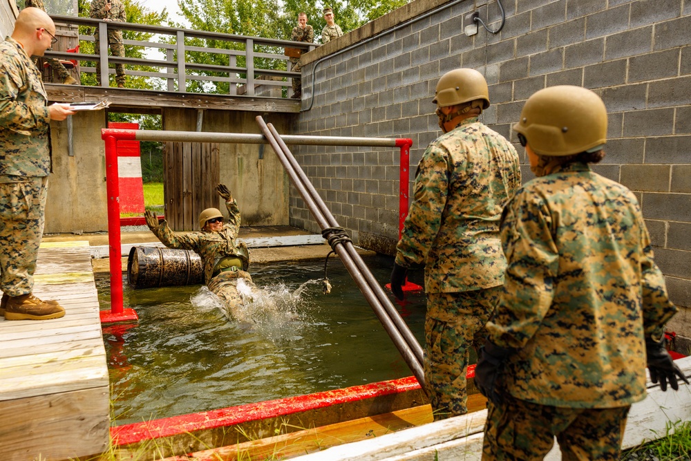 Military personnel in MARPAT camouflage uniforms and tan helmets are conducting a water obstacle course training exercise. One service member is submerged in a water pit with arms raised, while others assist using long poles and an evaluator records observations on a clipboard. Additional observers watch from an elevated platform in the background.