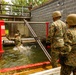 Military personnel in MARPAT camouflage uniforms and tan helmets are conducting a water obstacle course training exercise. One service member is submerged in a water pit with arms raised, while others assist using long poles and an evaluator records observations on a clipboard. Additional observers watch from an elevated platform in the background.