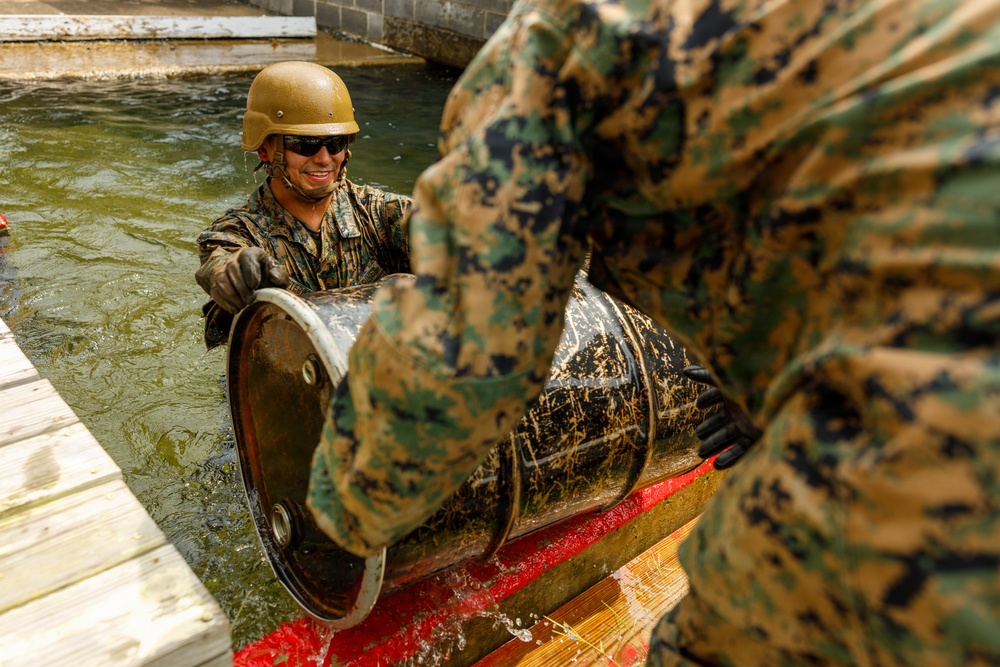 Two military personnel in woodland camouflage uniforms are working in water near a dock, handling a large metal barrel or drum. The soldier in focus is wearing a tan/coyote helmet, sunglasses, and tactical gloves while smiling. They appear to be engaged in a training exercise involving the movement or placement of metal drums in a water environment.
