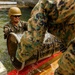 Two military personnel in woodland camouflage uniforms are working in water near a dock, handling a large metal barrel or drum. The soldier in focus is wearing a tan/coyote helmet, sunglasses, and tactical gloves while smiling. They appear to be engaged in a training exercise involving the movement or placement of metal drums in a water environment.