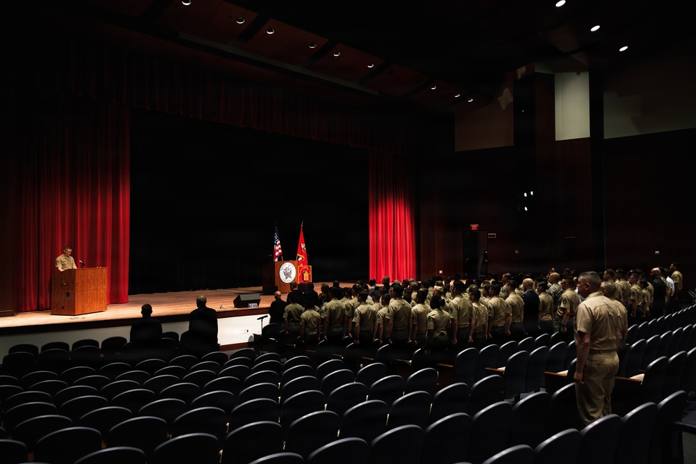 A military ceremony is taking place in a formal auditorium or theater setting. Personnel in tan/khaki uniforms are standing in formation facing a stage with a podium, flags, and red curtains. A speaker addresses the assembled group from a lectern on the left side of the stage.
