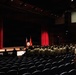 A military ceremony is taking place in a formal auditorium or theater setting. Personnel in tan/khaki uniforms are standing in formation facing a stage with a podium, flags, and red curtains. A speaker addresses the assembled group from a lectern on the left side of the stage.