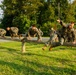 A group of military personnel in camouflage uniforms are navigating a wooden log obstacle course on a grassy field. Multiple individuals are visible vaulting over or traversing a horizontal log barrier supported by wooden posts. Additional personnel stand in the background observing the exercise.