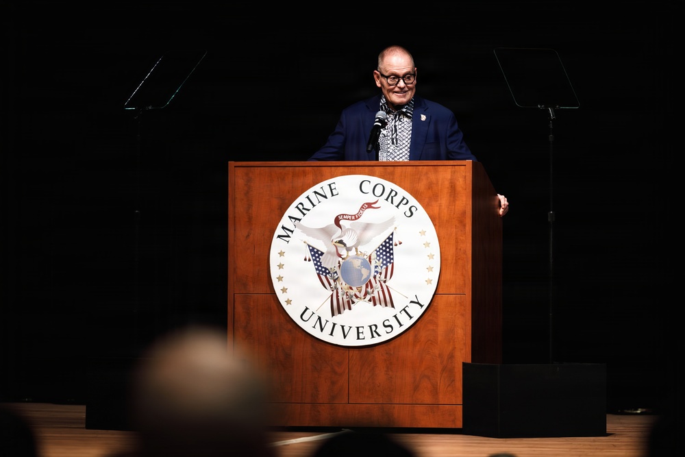A speaker stands at a wooden podium bearing the official seal of Marine Corps University, addressing an audience in a darkened auditorium or lecture hall. The individual is wearing a navy blue blazer with a patterned shirt and scarf, speaking into a microphone. The setting suggests a formal academic or institutional event hosted by Marine Corps University.