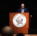 A speaker stands at a wooden podium bearing the official seal of Marine Corps University, addressing an audience in a darkened auditorium or lecture hall. The individual is wearing a navy blue blazer with a patterned shirt and scarf, speaking into a microphone. The setting suggests a formal academic or institutional event hosted by Marine Corps University.