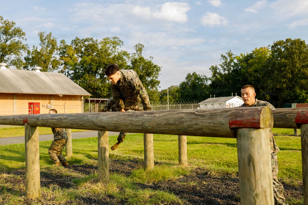 Military personnel in camouflage uniforms are navigating a wooden log obstacle course on a grassy training field. At least three individuals are visible, with one actively climbing over a large horizontal log beam supported by vertical wooden posts. The setting appears to be a military training facility during daylight hours with warm golden light suggesting early morning or late afternoon.