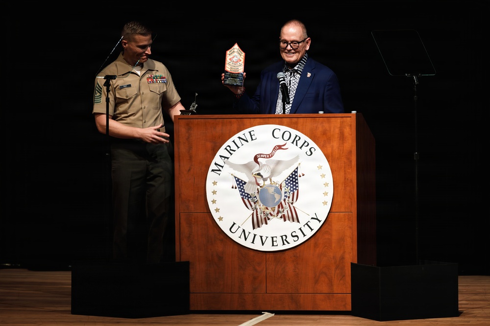 Two individuals are standing at a Marine Corps University podium during what appears to be an award ceremony. A civilian in a blue blazer is holding up a trophy/award while standing at the lectern, alongside a U.S. Marine in dress tan uniform. The setting appears to be a formal auditorium or stage event.