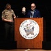 Two individuals are standing at a Marine Corps University podium during what appears to be an award ceremony. A civilian in a blue blazer is holding up a trophy/award while standing at the lectern, alongside a U.S. Marine in dress tan uniform. The setting appears to be a formal auditorium or stage event.